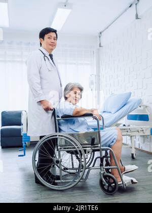 Paziente anziana asiatica vestita in azzurro, sorridente si siede felicemente in sedia a rotelle con medico gentilmente maschile in vestito bianco in piedi accanto a lei Foto Stock