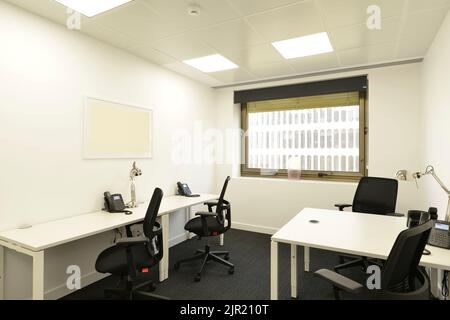 Armadio ufficio con un piccolo tavolo doppio bianco con telefoni, sedie girevoli nere, una finestra con vista e un pavimento in moquette grigio scuro Foto Stock