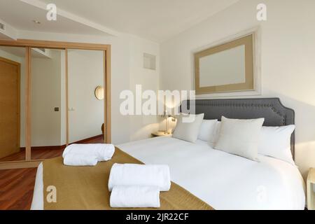 camera da letto con letto matrimoniale con asciugamani puliti, coperta di senape e armadio con grandi porte a specchio scorrevoli con bordi in legno di quercia e testiera rivestita grigia Foto Stock