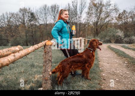 Giovane donna bella sport con il cane Irish Setter è a piedi lungo la strada della città, tenendo il caffè in tazza thermo tazza. Foto Stock