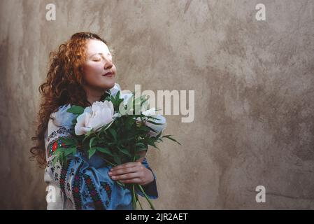 Ragazza in camicia ricamata bouquet di peonie bianche. Donna con lunghi capelli ricci rossi e occhi chiusi contro parete di fondo in cemento. Foto chiave scura. Giornata dell'indipendenza dell'Ucraina. Copia spazio. Messa a fuoco morbida Foto Stock