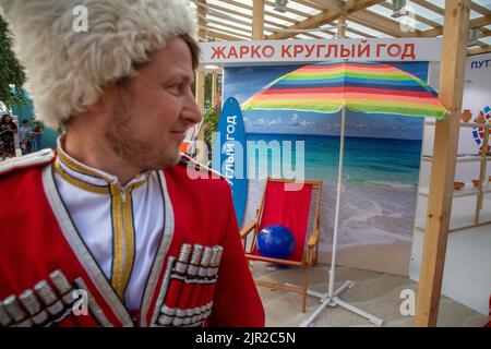 Mosca, Russia. 19th agosto 2022. Un uomo in costume Cossack allo stand della regione di Krasnodar e un'installazione pubblicitaria del turismo nazionale al V Festival della Società geografica Russa nel Parco Zaryadye nel centro di Mosca, Russia. Il banner recita: 'È caldo tutto l'anno' Foto Stock