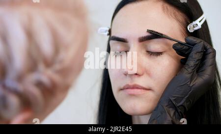 salone di bellezza. primo piano, un estetista in guanti di gomma nera, pettina delicatamente le sopracciglia del cliente con uno speciale pennello, pettine, forma le sopracciglia dopo la colorazione. Foto di alta qualità Foto Stock