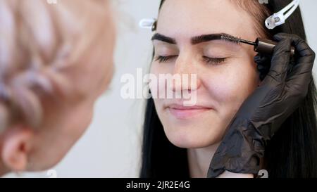 salone di bellezza. primo piano, un estetista in guanti di gomma nera, pettina delicatamente le sopracciglia del cliente con uno speciale pennello, pettine, forma le sopracciglia dopo la colorazione. Foto di alta qualità Foto Stock