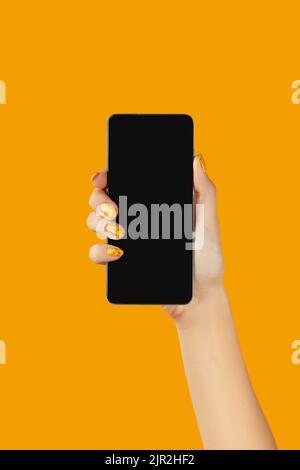 Donna ben curata che tiene lo smartphone nero con schermo vuoto su sfondo arancione Foto Stock
