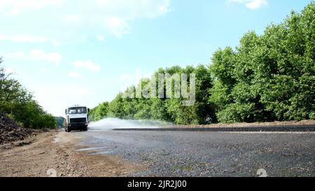 Riparazione di un'autostrada, lavori di costruzione stradale. Una speciale macchina irrigatrice spruzza acqua su una nuova pavimentazione in asfalto fresco, coprendo un lato del traffico. Foto Stock
