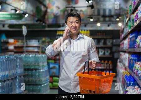 Un compratore maschio in un supermercato sceglie le merci e fa gli acquisti, un uomo asiatico parla allegramente sul telefono, consulta circa lo shopping Foto Stock