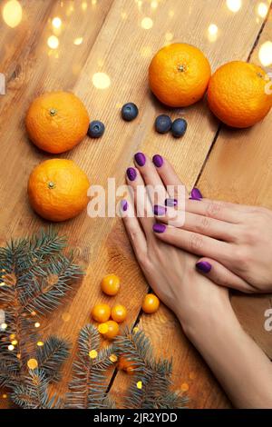 Mani delle donne con manicure borgogna sdraiato sul tavolo, vista dall'alto. Comfort domestico, tangerini, ramo di abete sullo sfondo delle luci di Natale. Foto di alta qualità Foto Stock