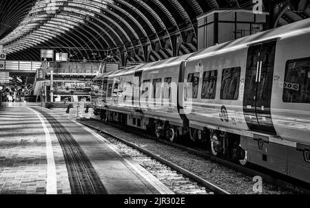 Accanto alla piattaforma della stazione ferroviaria si trova un treno. Ci sono passeggeri sullo sfondo e alcuni camminano attraverso un ponte pedonale. Una storica tettoia curva è Foto Stock