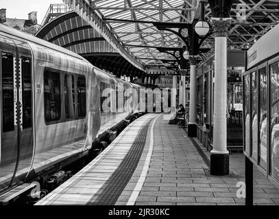 Accanto alla piattaforma della stazione ferroviaria si trova un treno. Sopra si trova uno storico baldacchino del 19th° secolo sorretto da colonne. Un passeggero siede su una panca e un adve Foto Stock