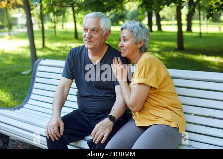 Abbastanza multi razziale anziani 60s coppia che riposano sedersi sulla panca nel parco dopo una passeggiata, mattina a piedi sorridendo godere parlare rilassante all'aperto nella soleggiata giornata estiva Foto Stock