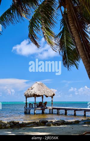 Un luogo idilliaco su Ambergris Caye, San Pedro, Belize. Due sedie sotto una palapa che si affaccia sull'acqua blu, con palme e una spiaggia in primo piano. Foto Stock