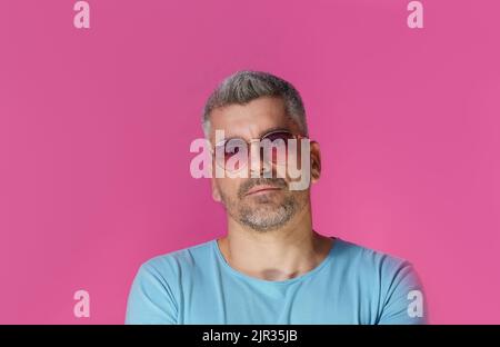 Bell'uomo 30s 40s che indossa occhiali da sole rosa e t-shirt blu con un look aggressivo e concentrato isolato su sfondo rosa. Un uomo maturo dai capelli grigi girato in studio. Foto Stock