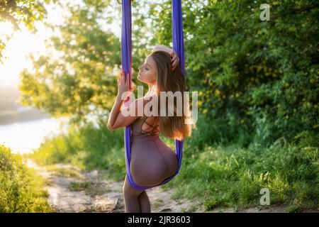 Una giovane ginnastica femminile è impegnata nello yoga aereo, utilizzando una combinazione di pose yoga tradizionali, pilates e danza utilizzando un'amaca al tramonto in natura Foto Stock