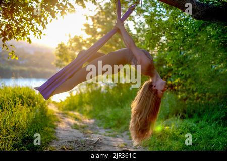 Una giovane ginnastica femminile è impegnata nello yoga aereo, utilizzando una combinazione di pose yoga tradizionali, pilates e danza utilizzando un'amaca al tramonto in natura Foto Stock