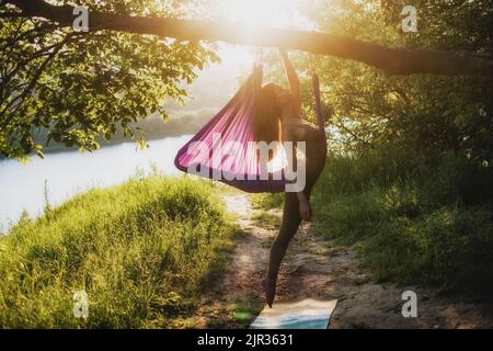 Una giovane ginnastica femminile è impegnata nello yoga aereo, utilizzando una combinazione di pose yoga tradizionali, pilates e danza utilizzando un'amaca al tramonto in natura Foto Stock