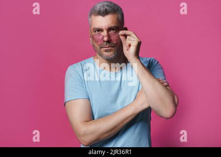 Serio grigio capelli bell'uomo in casual isolato su sfondo rosa. Bell'uomo di mezza età in piedi con le braccia ripiegate abbassò gli occhiali da sole guardando la fotocamera indossando una t-shirt blu. Foto Stock