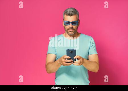 Uomo bello concentrato e penoso 30s 40s che guarda lo smartphone in mano indossando occhiali da sole blu e t-shirt isolata su sfondo rosa. Uomo mucho in t-shirt blu con scatto da studio per telefono. Foto Stock