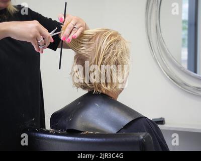 Un parrucchiere con un pettine e forbici che tagliano i capelli di un cliente biondo al salone di bellezza Foto Stock