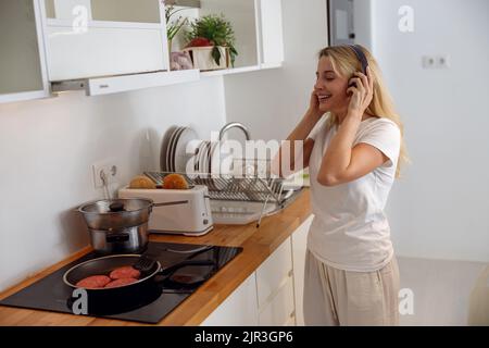 Bionda bella donna in cuffie ascoltare musica e cucinare in cucina. Foto Stock