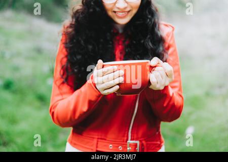 Bruna donna riccia, indossando una giacca di pelle rossa, e tenendo una tazza rossa con caffè caldo in natura. Foto Stock
