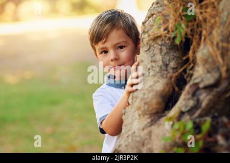 Nascondere e sbirciare un ragazzino felice che gioca accanto ad un albero nel parco. Foto Stock