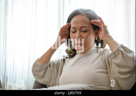 Una donna di età asiatica malata soffre di mal di testa o emicrania mentre si rilassa nel suo confortevole soggiorno. Foto Stock