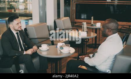 Due uomini d'affari multietnici seduti, sorridenti e che discutono della loro partenza in un arioso caffè durante l'ora di pranzo. Le tazze di caffè sono sul loro tavolo Foto Stock