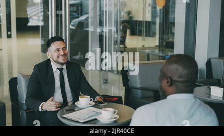 Allegro uomo d'affari caucasico bearded in abiti formali sorridendo, gesticulating, bevendo caffè e discutendo il rapporto finanziario di startup con il suo socio afro-americano in caffè moderno Foto Stock