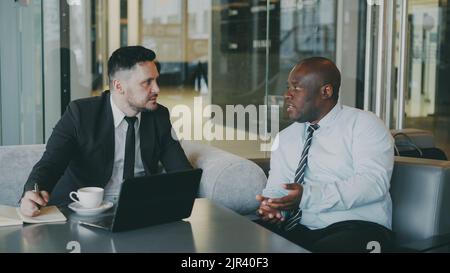 Due uomini d'affari multirazziali sicuri che discutono circa la collaborazione durante l'incontro in caffè moderno vetroso Foto Stock