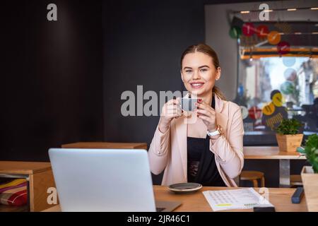 Donna sorridente seduta in caffetteria con tazza di caffè e lavoro sul computer portatile Foto Stock