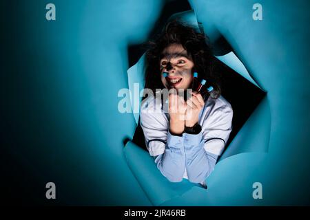 MAD scienziato con capelli capienti e provette di vetro pieno che grigning terribile alla macchina fotografica mentre su sfondo blu. Chimico maniaco sciolto con vassoio con provette riempite di composti tossici Foto Stock