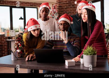 Team diversificato di colleghi che utilizzano il notebook in ufficio durante le festività natalizie, che lavorano alle startup durante le festività natalizie. Fare lavoro di squadra in un luogo di lavoro decorato con ornamenti di natale. Foto Stock