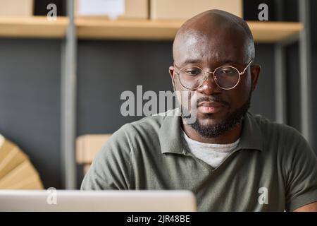 Uomo serio africano in occhiali che lavorano online utilizzando il laptop in magazzino Foto Stock