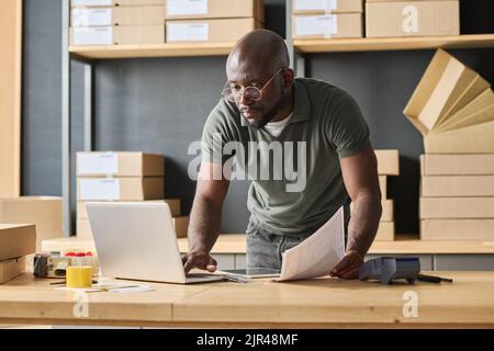 Serio uomo africano concentrandosi sul suo lavoro sul laptop a tavolo, ha digitato i dati dei pacchi durante il suo lavoro in magazzino Foto Stock