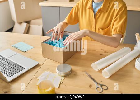 Primo piano di una donna che imballa il pacco per la consegna seduto al tavolo di fronte al portatile per lavorare con gli ordini online Foto Stock