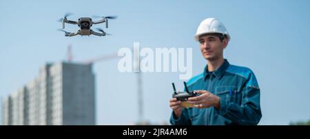 Un uomo in un casco e tute controlla un drone in un cantiere. Il costruttore esegue la supervisione tecnica. Foto Stock