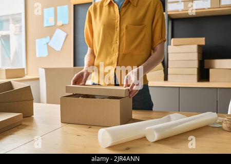 Primo piano di una giovane lavoratrice che chiude la scatola di cartone e finisce con gli ordini di imballaggio al tavolo in magazzino Foto Stock