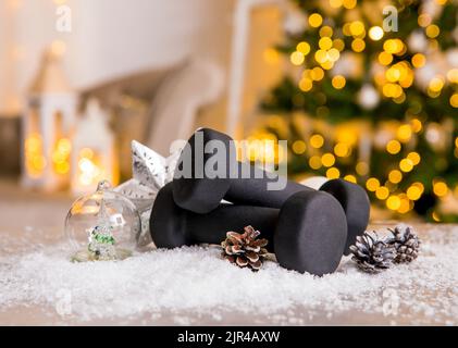 Stile di vita sano durante il periodo di festa di Natale concetto. Manubri neri con ornamenti natalizi e neve offuscata albero di Natale sullo sfondo. Foto Stock