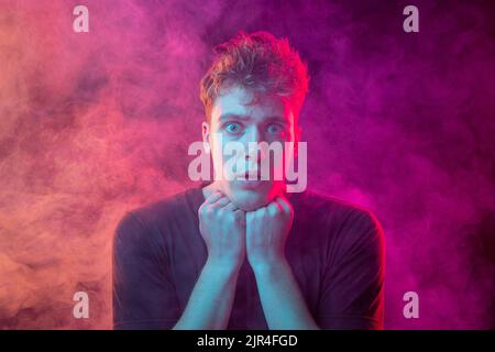 Giovane uomo avvolto nel fumo. Modello che guarda la telecamera isolata su sfondo magenta con nuvole di fumo. Concetto di salute mentale, arte, emozioni Foto Stock