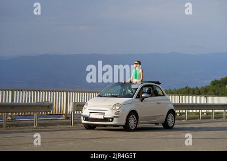 Donna viaggiatore gode di paesaggi rocciosi sopra le nuvole in alta montagna da una piccola auto convertibile. Foto Stock