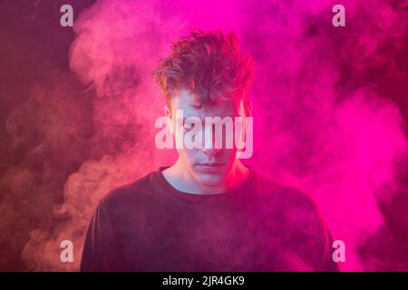 Giovane uomo avvolto nel fumo. Modello che guarda la telecamera isolata su sfondo magenta con nuvole di fumo. Concetto di salute mentale, arte, emozioni Foto Stock