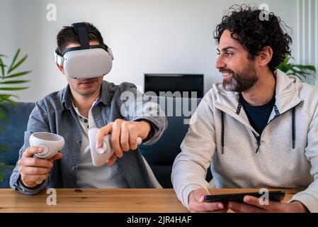 Gli uomini caucasici godendo di occhiali di realtà virtuale mentre si siedono al tavolo in casa. Due happy ragazzi che gesturano e utilizzano il visore vr o 3D occhiali da gioco Foto Stock