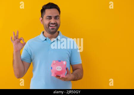 Giovane uomo allegro che mostra il segno OK mentre si trova in piedi con una banca piggy in mano Foto Stock