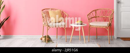 Sedie in vimini con cuscini, tavolo e ciotola con frutta vicino alla parete rosa in camera Foto Stock