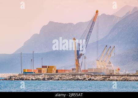 Costruzione di gru industriali e di lavoro nel porto di carico della città di Antalya con navi sullo sfondo delle montagne al tramonto. Settore delle spedizioni marittime Foto Stock