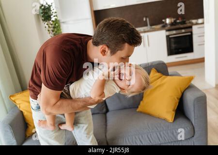 Giovane padre coccola il figlio del bambino dopo essere tornato a casa dal lavoro, abbracciandolo, solletico tenendolo in mano sullo sfondo della cucina e sul divano grigio nel soggiorno. Felice infanzia spensierata, paternità Foto Stock