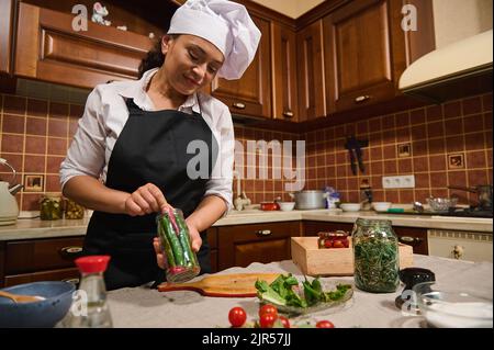 Casalinga in cappellino bianco chef e grembiule nero, si riempie di lattina con peperoncino fresco, facendo sottaceti e prelibatezze marinate Foto Stock