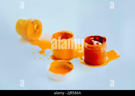 Su un tavolo bianco ci sono due vasi di gouache arancione e rosso, e un vaso giallo giace sul suo lato e la vernice gialla scorre fuori da esso. Confusione creativa. Insp Foto Stock