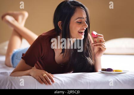 Felice giovane donna che mangia i macaroon mentre sdraiata sul letto Foto Stock